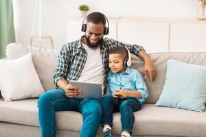 Young African Father And His Kid Boy Wearing Headphones Using Tablet Computer Together Watching Movie Online On Weekend Sitting On Sofa At Home. Weekend With Daddy. Family Leisure And Gadgets photo
