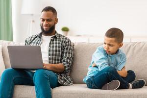 Unhappy Black Kid Boy Sitting Near Reluctant Indifferent Father While He Using Laptop Not Paying Attention To Offended Little Son Sitting On Sofa At Home. Family Problems, Bad Relationship With Dad photo