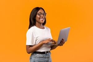 Happy african american young woman with glasses holding newest laptop and smiling, orange studio background, panorama with copy space. Cheerful black lady student using notebook, studying online photo