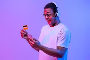 Cashback Concept. Surprised black guy in wireless headphones holding smartphone and credit card, emotionally reacting to online bonus, standing in neon light over purple background, free space photo