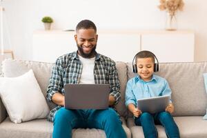 Black Father And Little Boy Using Computers Together Browsing Internet On Laptop And Digital Tablet Wearing Wireless Headphones Sitting On Sofa At Home. Family Gadgets Concept photo