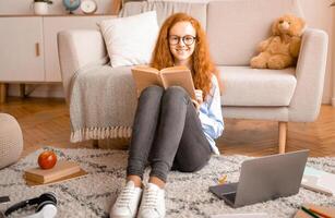Education Concept. Full length portrait of cute smiling red-haired girl wearing glasses, sitting on the floor carpet with laptop, reading paper book, looking at camera. Teen resting at home photo
