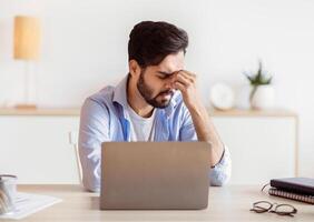 Desperate Eastern Freelancer Guy Sitting At Desk With Laptop, Suffering Lack Of Inspiration Or Feeling Headache, Stressed Young Arab Businessman Having Problems At Work, Free Space photo