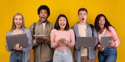 Gossip, shocking news, study with online technology and teamwork. Surprised teens multiracial students learning, using digital gadgets, isolated on orange background, copy space, studio shot photo