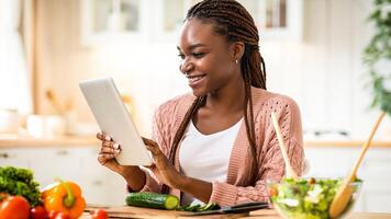 Online Recipe. Cheerful african american woman using digital tablet in kitchen while cooking healthy lunch, smiling black female checking ingredients in internet or reading culinary blog, free space photo