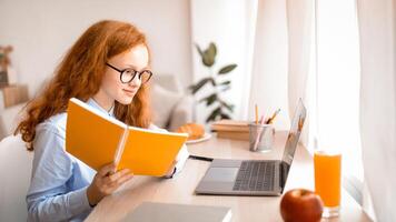 Doing Homework. Portrait of pretty young teenage schoolgirl wearing glasses, holding notebook and using laptop computer, sitting at table, studying and leraning in her bedroom photo
