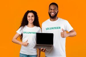 Volunteering Website. Two Happy Activists Showing Laptop Blank Screen And Like Gesture Standing Over Yellow Background In Studio. Couple Of Diverse Volunteers Hold Computer With Empty Display. Mockup photo