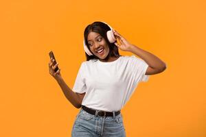 Joyful young african american lady listening to music over orange studio background, using wireless headphones and smartphone. Smiling black woman enjoying newest stereo headset and mobile music app photo