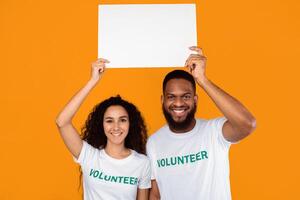 Couple Of Multiethnic Volunteers Holding Empty White Paper Board Above Heads Advertising Your Text Posing Over Yellow Studio Background. Volunteering And Charity Banner Advertisement. Mockup photo