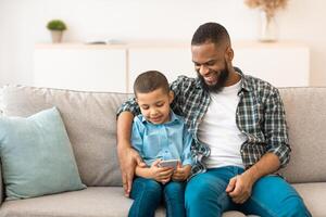 Black Boy And His Dad Using Mobile Phone Playing Games Online Sitting Together On Couch And Browsing Internet In Living Room Indoor. New Application For Smartphone, Family And Gadgets Concept photo