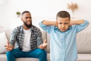 Black Boy Covering Ears While Angry Father Shouting At Him Standing At Home, Looking At Camera. Domestic Violence And Unhappy Childhood, Family Problem And Bad Relationship. Selective Focus photo