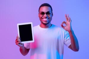 Happy African American Guy In Neon Lighting Holding Digital Tablet With Black Screen And Showing Ok Gesture, Recommending New Website Or App While Standing Over Purple Background, Mockup Image photo