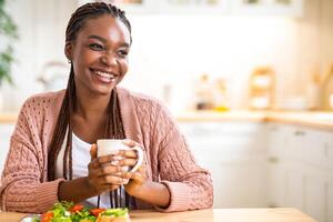 Start Of The Day. Happy Black Woman Eating Breakfast And Drinking Morning Coffee In Kitchen, Portrait Of Dreamy Young African American Lady Sitting At Table With Cup Of Hot Drink In Hands And Smiling photo