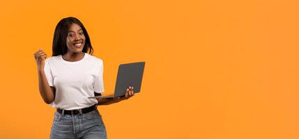 Smiling young black woman in white t-shirt holding laptop and raising fist up over orange studio background, catch nice discounts for online shopping or celebrating success in gambling, copy space photo