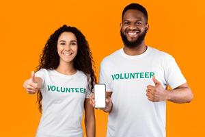 Charity Application. Two Cheerful Multicultural Volunteers Showing Phone Empty Screen Gesturing Thumbs Up Posing Standing Over Yellow Studio Background. We Approve This App Concept. Mockup photo