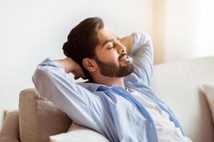 Relax Time. Portrait Of Handsome Arab Man Leaning Back On Couch With Closed Eyes, Keeping Hands Behind Head, Eastern Guy Resting On Comfortable Sofa In Living Room, Side View With Copy Space, Closeup photo