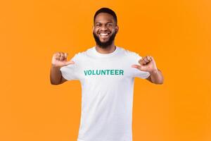 I'm a volunteer. Cheerful black activist guy pointing fingers at himself and volunteer inscription on his t-shirt posing smiling to camera on yellow studio background. Volunteering work concept photo