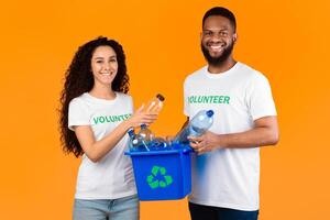 Two Diverse Volunteers Holding Box With Recycle Symbol And Plastic Bottles Posing Over Yellow Studio Background, Smiling To Camera. Junk Disposal, Recycling And Waste Sorting Concept photo
