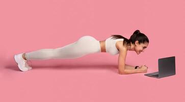 Training Concept. Full length portrait of sporty smiling woman doing plank while watching online tutorial on laptop computer, exercising on the floor, isolated over pink studio background photo