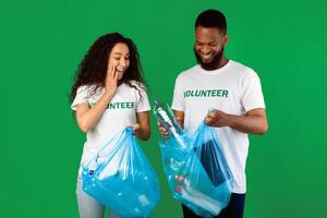 Sort Your Waste. Two Multiracial Volunteers Couple Putting Used Plastic Bottle To Garbage Bag Posing Standing In Studio Over Green Background. Keep Environment Clean, Ecology Concept photo