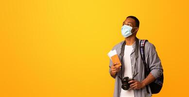 New Travel Rules. Young Black Tourist Man In Medical Face Mask Looking Aside At Copy Space, African Guy With Passport And Photo Camera Ready For Trip During Coronavirus Pandemic, Yellow Background