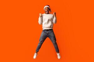 Overjoyed Black Guy In Santa Hat Celebrating Success, Jumping Over Orange Background With Raised Fists, Euphoris African American Man Enjoying Christmas Offers And Seasonal Sales, Full Length Shot photo