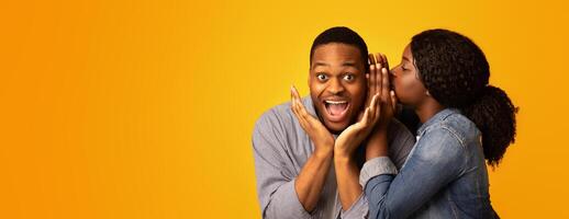 Gossips Concept. Black Woman Sharing Secret With Her Excited Boyfriend, Surprised African American Guy Opening Mouth In Amazement While Standing Together Over Yellow Studio Background, Free Space photo