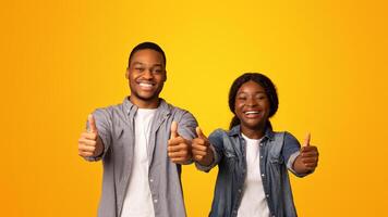 We Recommend. Portrait of cheerful african american couple gesturing thumbs up at camera, expressing approval, joyful black man and woman standing isolated on yellow background, copy space photo