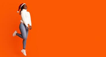 Christmas Mood. Portrait Of Happy Jumping Black Lady In Santa Hat Over Orange Background, Cheerful African American Woman Having Fun, Enjoying Xmas And New Year Holidays, Full Length Shot, Copy Space photo