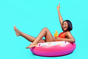 Cool black lady sitting on inflatable ring, having fun, enjoying summer cocktail on blue background, full length. Cheerful young woman having beach party, chilling and screaming in excitement photo
