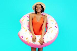 Portrait of joyful millennial black lady in swimsuit posing with inflatable ring on blue studio background. Positive young woman enjoying summertime, having fun at beach party or tropical vacation photo