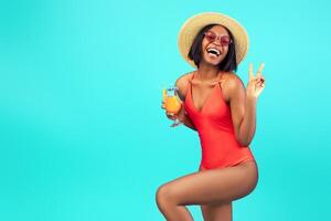 Happy black woman in swimsuit enjoying exotic summer cocktail and showing peace sign at camera, posing over blue studio background. Playful African American lady having fun on beach vacation photo