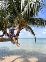 Man in sunglasses sits on a slanted palm tree holding a coconut in his hands. In the background is the turquoise sea, blue sky and tropical landscape of Phu Quoc Island in Vietnam. photo