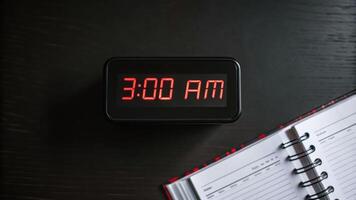 Digital clock 3AM red LED dark background planner open notebook time display early morning black surface spiral binding schedule organization photo