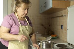 A senior woman in an apron is using a knife to add butter to a saucepan on the stove. photo