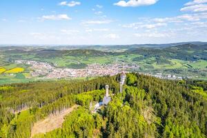 Aerial view captures the Svatobor Lookout Tower surrounded by lush green forests, overlooking the town of Susice nestled in the valley below on a clear day. photo