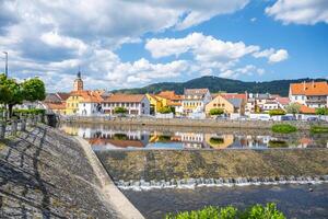The tranquil waters of the Otava River reflect colorful buildings in Susice, Czechia. Visitors enjoy a sunny day with a backdrop of green hills and blue skies. photo