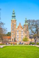 Visitors stroll through the gardens of Rosenborg Castle, a stunning renaissance structure in Copenhagen, Denmark. The clear blue sky enhances the beauty of the historic architecture. photo