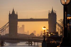 Sunrise casts a warm glow on Tower Bridge as it stands majestically over the River Thames. The surrounding city skyline fades into the background, creating a stunning silhouette. photo