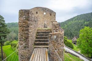Visitors walk across the bridge of Velhartice Castle, surrounded by lush greenery and steep hills. The ancient stone structure offers a glimpse into history and stunning views. photo
