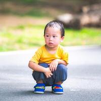 Asian boy's head is soaked in sweat from the hot and humid weather. Child squats in the middle of the road to rest while walking. The 2-year-old kid is wearing a dark yellow shirt. photo