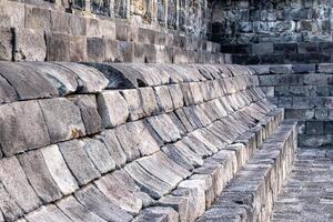 Stone seating structure at Borobudur Temple with worn, curved blocks and ancient carvings above. photo