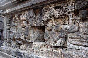 Ancient stone relief showing people, trees, and intricate carvings on a temple wall. photo