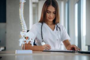 Writing in the document, spine model is on the table. Professional female doctor in the clinic indoors photo