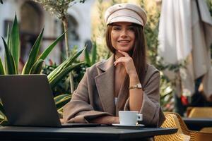 Looking, at the camera, sitting. Beautiful young woman is outdoors of the restaurant by the table photo