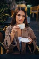 Looking, at ,,the camera, sitting. Beautiful young woman is outdoors of the restaurant by the table photo