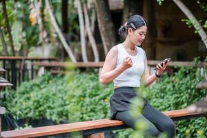 A young woman in athletic clothing sits on a wooden railing, focused on her smartphone, surrounded by vibrant greenery. photo