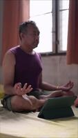 Meditation concept. A man is sitting on a bed with a tablet in front of him. He is wearing a purple shirt and green shorts. Scene is calm and peaceful, as the man is practicing yoga and meditating video
