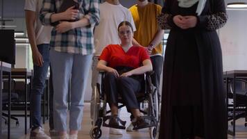 A diverse group of young business people congregates within a modern startup's glass-enclosed office, featuring inclusivity with a person in a wheelchair, an African American young man , and a hijab video