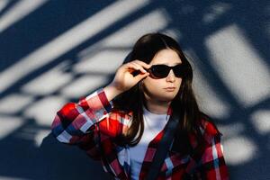 Young student adjusting sunglasses while leaning against wall with shadows. photo